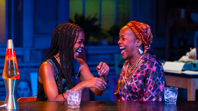 two actors laughing at a table on stage