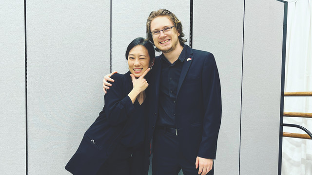 Two people, identified in the accompanying caption, stand side by side, smiling warmly. Both are dressed in black attire, and they stand against a light-colored background, exuding a sense of camaraderie and joy.
