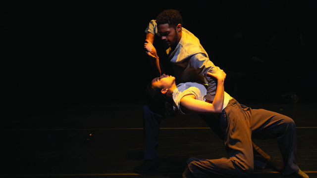 Two dancers, named in the accompanying caption, perform an expressive and intimate dance on stage under dim lighting. The focus is on their intense connection and the fluidity of their movements, creating a powerful and emotional performance.